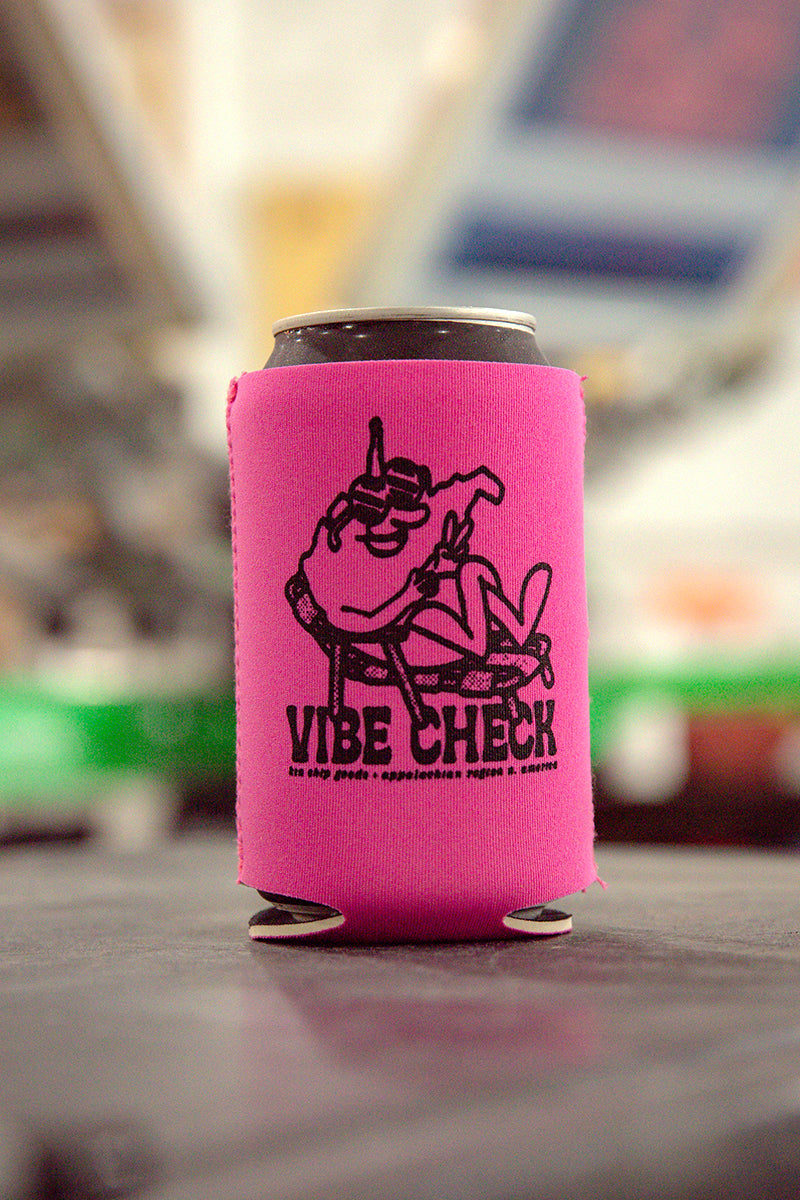 Kin Ship Goods “Vibe Check” koozie in bright pink with lounging West Virginia figure and black peace sign graphic.
