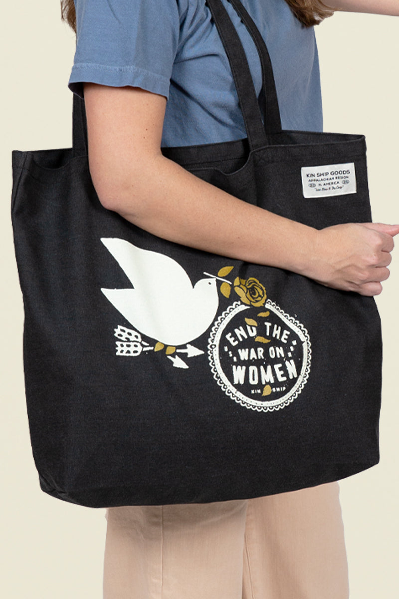 Kin Ship Goods black tote bag with white dove, gold rose, and 'End the War on Women' message in bold black and white circle.